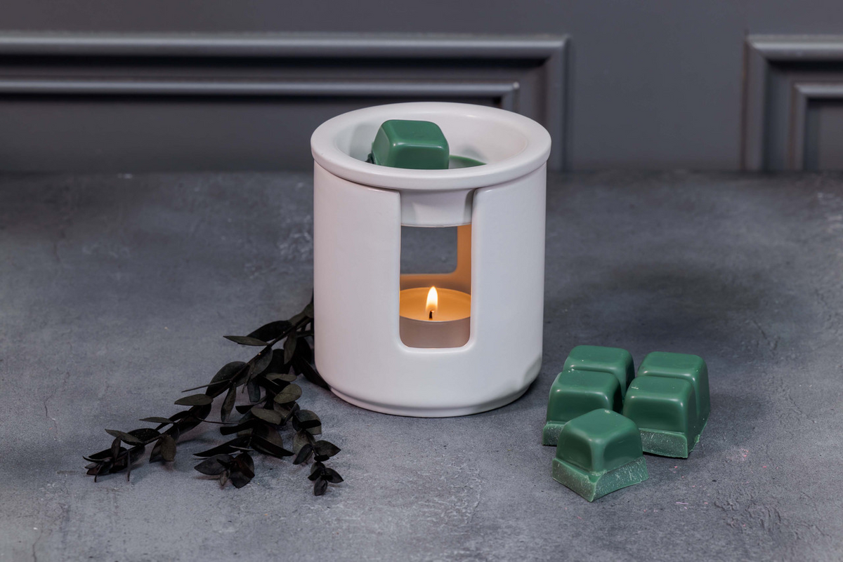 White wax warmer with a lit candle and green wax melts on a gray surface.