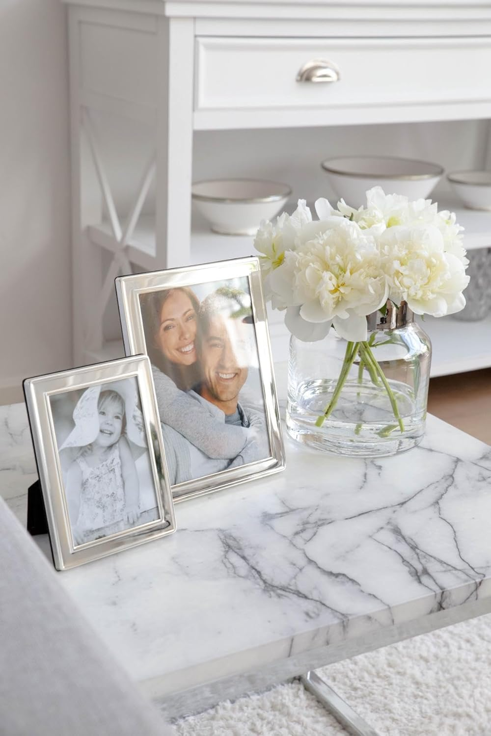 Two silver photo frames with family portraits on a marble surface next to a clear vase with white flowers.